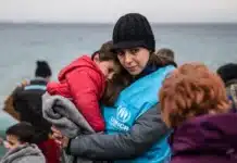 Kvinde holder barn med havet i baggrunden. De er omgivet af mennesker.