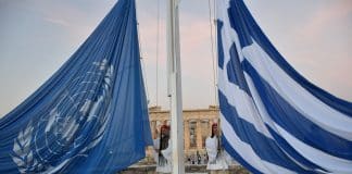 Δύο τσολιάδες στην Ακρόπολη ανυψώνουν τη σημαία της Ελλάδας και του ΟΗΕ, με φόντο τον Παρθενώνα, σε μια τελετουργική στιγμή που συμβολίζει τη διεθνή συνεργασία και την ελληνική πολιτιστική κληρονομιά.