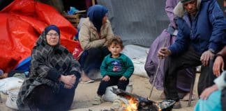 Τρεις ηλικιωμένοι και ένα παιδί καθισμένοι γύρω από μια μικρή φωτιά, αντανακλώντας τις δυσκολίες των εκτοπισμένων Παλαιστινίων στη Γάζα.