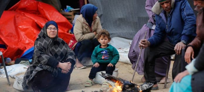 Τρεις ηλικιωμένοι και ένα παιδί καθισμένοι γύρω από μια μικρή φωτιά, αντανακλώντας τις δυσκολίες των εκτοπισμένων Παλαιστινίων στη Γάζα.