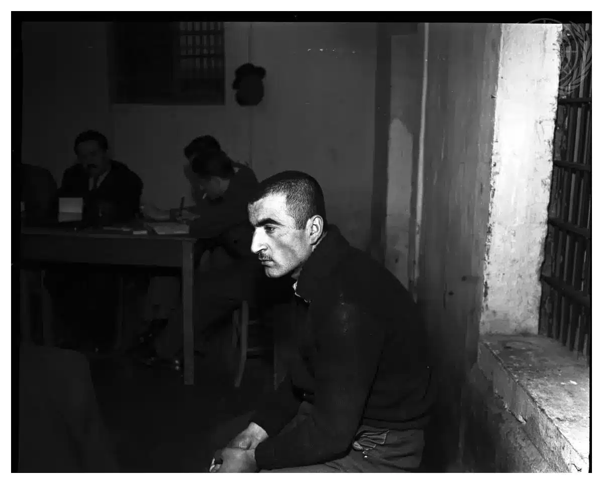 14 Νεαρός άνδρας καθισμένος δίπλα σε ένα παράθυρο εξετάζεται, σκυθρωπός, από επιτροπή.