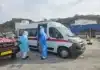 Medische evacuatie in een haven in Kaapverdië op 6 mei 2026. Drie hulpverleners in volledige blauwe en witte beschermende pakken (PPE) staan bij de open schuifdeur van een witte ambulance. Eén hulpverlener houdt een brancard vast, terwijl een ander in de ambulance stapt. Links staat een rood voertuig met de tekst 'AMBULANCE' in spiegelbeeld op de motorkap. Op de achtergrond zijn een havenkade, vissersboten en gebouwen tegen een heuvel zichtbaar.