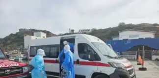 Wereldgezondheidsorganisatie: hantavirus-respons op besmettingen van cruiseschip Medische evacuatie in een haven in Kaapverdië op 6 mei 2026. Drie hulpverleners in volledige blauwe en witte beschermende pakken (PPE) staan bij de open schuifdeur van een witte ambulance. Eén hulpverlener houdt een brancard vast, terwijl een ander in de ambulance stapt. Links staat een rood voertuig met de tekst 'AMBULANCE' in spiegelbeeld op de motorkap. Op de achtergrond zijn een havenkade, vissersboten en gebouwen tegen een heuvel zichtbaar.