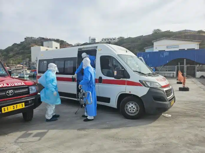 Medische evacuatie in een haven in Kaapverdië op 6 mei 2026. Drie hulpverleners in volledige blauwe en witte beschermende pakken (PPE) staan bij de open schuifdeur van een witte ambulance. Eén hulpverlener houdt een brancard vast, terwijl een ander in de ambulance stapt. Links staat een rood voertuig met de tekst 'AMBULANCE' in spiegelbeeld op de motorkap. Op de achtergrond zijn een havenkade, vissersboten en gebouwen tegen een heuvel zichtbaar.