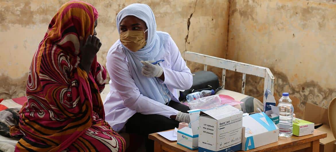 En kvinna med mask som sitter vid ett vaccinationsbord.