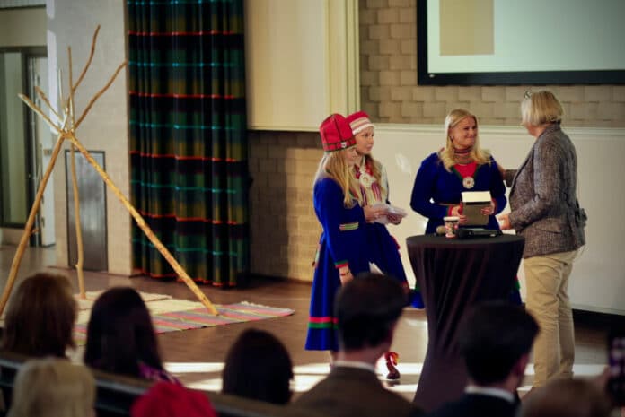 Två kvinnor i traditionell samisk klädsel överräcker en bok till en tredje kvinna på en scen